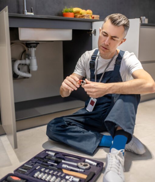 Reflections. Middle-aged man in white tshirt and blue overalls with badge sitting on floor looking at plumbing piece in hands near open set of tools in kitchen